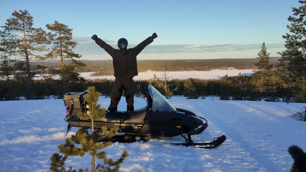Snowmobile gift card: a happy rider standing on a snowmobile above a frozen lake in Swedish Lapland